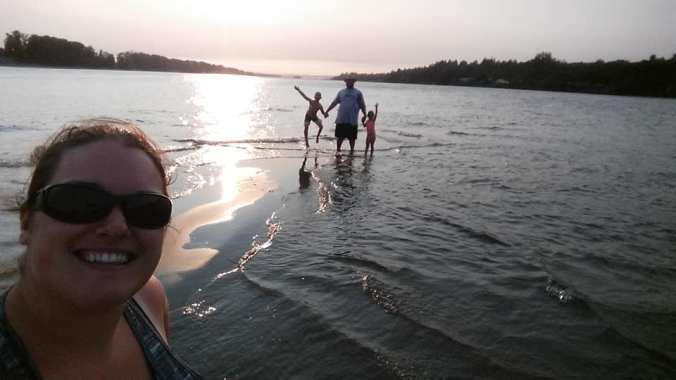 mom smiling in the foreground of a selfie with dad and two kids behind silhouetted against the sunset, standing and jumping in the water