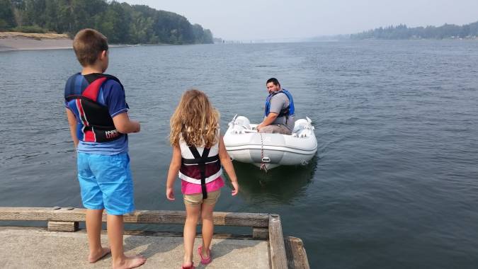 A boy and a girl standing on a dock watching their father approach in an inflatable dinghy, and island with trees in the background