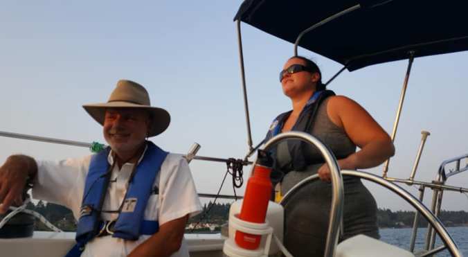 A woman at the helm of a sailboat named Mosaic, looking forward on the boat, with an instructor sitting in front of her smiling at the camera