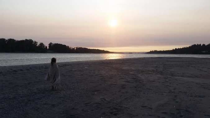 Young girl stands on a beach wrapped in a towel in a regal pose watching the sunset