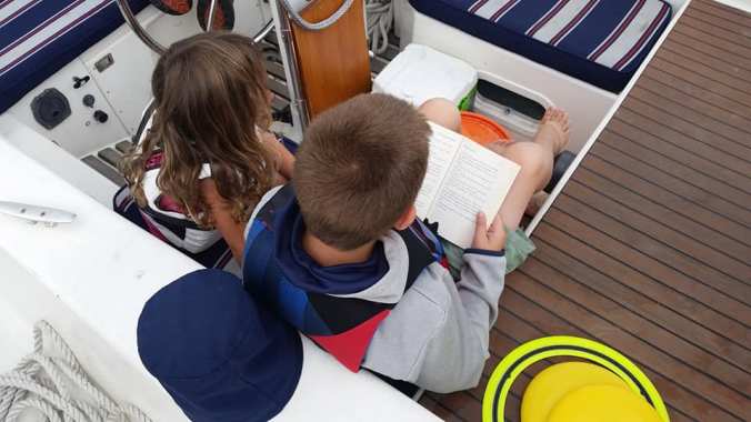 Boy sits in the cockpit of a sailboat next to a younger girl reading a book to her.