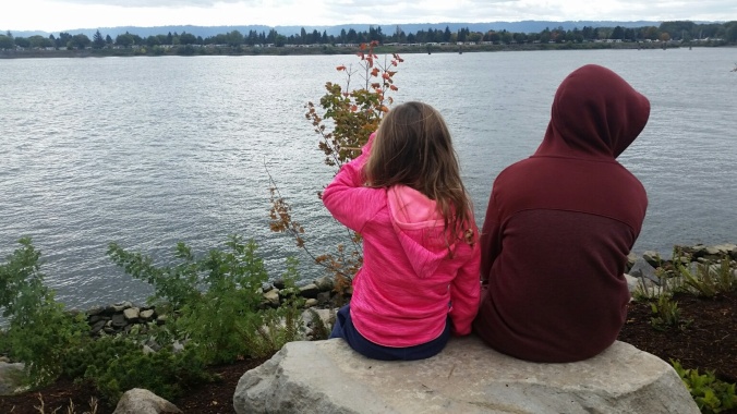 A boy and a girl sit side by side on a large rock overlooking the Columbia River.