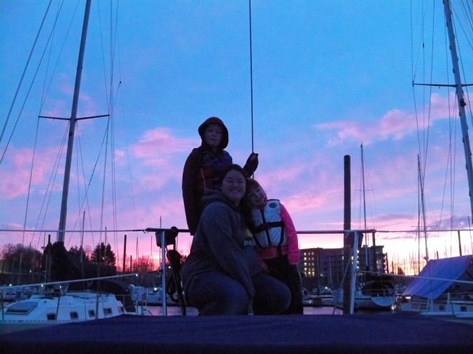 A woman and her two kids grab a photo on the bow of their sailboat, their home, with a beautiful March Sunset in Portland Oregon in the background