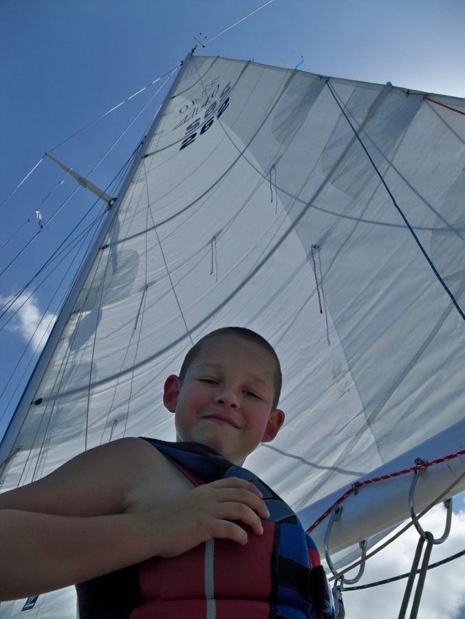 Our son Evan smiles at the camera with the large mainsail of our Fuji 40 sailboat rising into the sky behind him